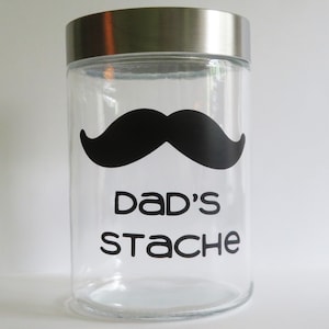 May include: A clear glass jar with a silver lid. The jar has a black moustache and the text "Dad's Stache" on the front.