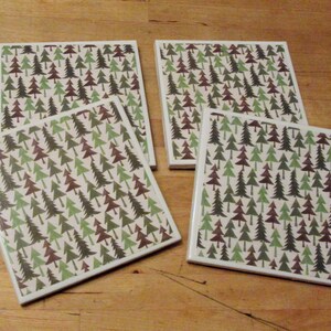 May include: Four square ceramic coasters with a repeating pattern of green and brown pine trees on a cream background. The coasters have white edges and are arranged on a wooden surface.
