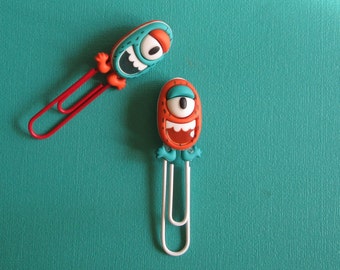 May include: Two colorful monster-shaped bookmark clips. One is blue and orange with a single eye and a wide smile. The other is red and orange with two eyes and a wide smile.