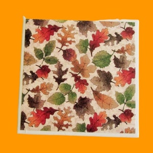 May include: Square coaster featuring a repeating pattern of autumn leaves in shades of red, orange, green, and brown. The leaves are set against a cream-colored background with a textured appearance. The coaster has a white border.