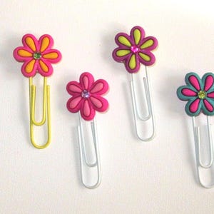 May include: Four colorful flower-shaped paper clips. The paper clips are yellow, pink, green, and blue. Each paper clip has a flower design with a rhinestone in the center.