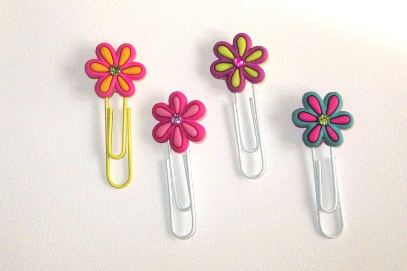 May include: Four colorful flower-shaped paper clips. The paper clips are yellow, pink, green, and blue. Each paper clip has a flower design with a rhinestone in the center.