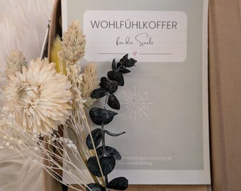 May include: A brown cardboard box with a white card inside. The card has the text "WOHLFÜHLKOFFER für die Seele" and a small pink heart. The box is filled with dried flowers and eucalyptus.