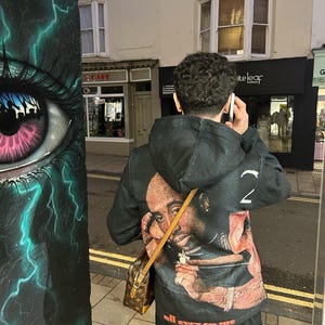 May include: A person wearing a black hoodie with a graphic print of a man's face and the text "all eyez on me". The person is holding a phone to their ear and has a brown leather bag slung over their shoulder.