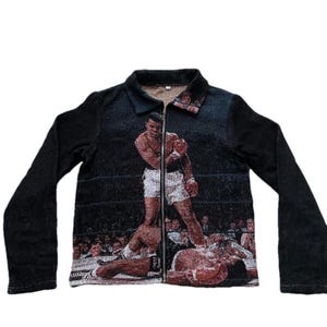 Puede incluir: Una chaqueta negra con cremallera que tiene una imagen tejida de un boxeador en un ring de boxeo. El boxeador lleva pantalones cortos blancos y guantes de boxeo negros.
