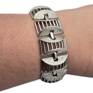 May include: Silver-toned bracelet with a geometric design. The bracelet features rectangular links with a half-circle design on each end, connected by a central bar. The links have vertical lines, creating a textured appearance. The bracelet is worn on a wrist.
