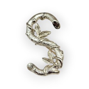 May include: Silver-toned brooch in the shape of the letter "S". The brooch is designed to resemble bamboo stalks with leaves. The pin is a decorative accessory.