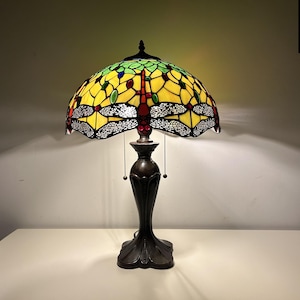 May include: A stained glass dragonfly lamp with a yellow and green shade. The lamp has a dark bronze base and a pull chain. The dragonfly design is visible on the shade. The lamp is on a white surface.