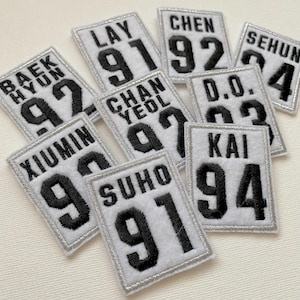 May include: Nine white fabric patches with black embroidered numbers and names. The patches include the names Baekhyun, Lay, Chen, Chanyeol, D.O., Xiumin, Suho, Kai, and Sehun.