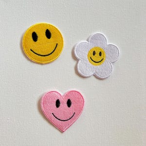 Peut inclure: Trois patchs brodés à repasser avec des visages souriants. Un cercle jaune, une fleur blanche et un cœur rose.