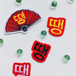 May include: Four red and white embroidered patches with the text "CCH" in yellow and black lettering. The patches are arranged on a white background with green marbles scattered around them. One patch is a red and white fan.