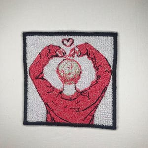 May include: Embroidered patch featuring a person making a heart shape with their hands, with a small heart above their head. The person is wearing a red sweater.