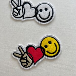 Peut inclure: Deux patchs brodés à repasser. Le premier patch est blanc avec un contour noir et représente un signe de paix, un cœur et un visage souriant. Le deuxième patch est rouge, jaune et blanc avec un signe de paix, un cœur et un visage souriant.
