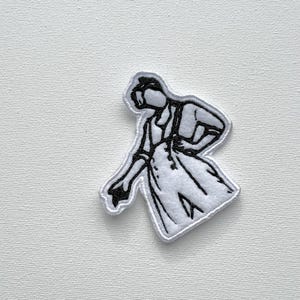 May include: White embroidered iron-on patch of a person in a white jumpsuit with a black outline. The person is in a dancing pose with one arm raised and the other arm bent at the elbow.