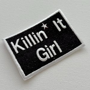Puede incluir: Parche rectangular negro con texto bordado blanco «Killin' It Girl» y una estrella. El parche tiene un borde blanco, ideal para ropa o accesorios.
