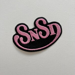 May include: A black and pink embroidered patch with the text "SNSD" in a cursive font.