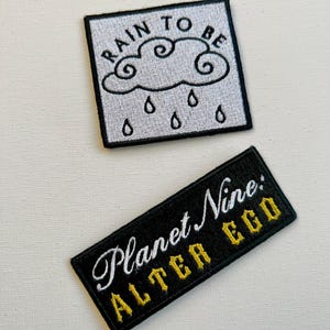 May include: Two embroidered patches. The first patch is white with black embroidery of a cloud with rain drops and the text "RAIN TO BE". The second patch is black with white embroidery of the text "Planet Nine: ALTER EGO" in gold.