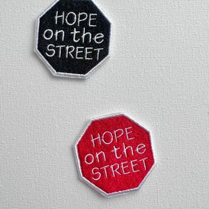 Könnte beinhalten: Zwei gestickte Patches in Form eines Stoppschildes. Der obere Patch ist schwarz mit weißem Text, der "HOPE on the STREET" lautet. Der untere Patch ist rot mit weißem Text, der "HOPE on the STREET" lautet.