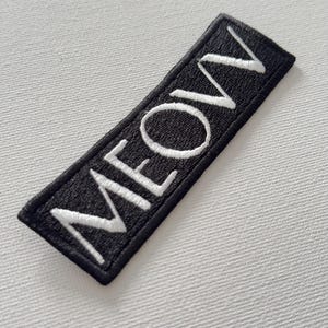 May include: Black fabric patch with white embroidered text that reads "MEOW".