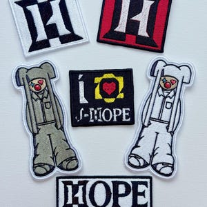 Könnte beinhalten: Eine Sammlung von gestickten Aufnähern mit verschiedenen Designs. Enthält Aufnäher mit dem Buchstaben "H" in Schwarz und Weiß sowie Rot und Weiß. Außerdem Aufnäher mit dem Text "i ♡ J-HOPE" und "HOPE ON THE STAGE". Zwei Aufnäher zeigen eine Cartoonfigur.