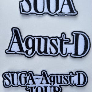 Puede incluir: Tres parches bordados en blanco y negro con el texto "SUGA", "Agust D" y "SUGA Agust D TOUR".