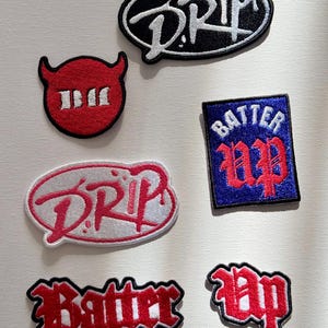 Peut inclure: Six patchs brodés thermocollants. Deux patchs sont rouges avec du texte blanc qui dit "Batter Up". Un patch est rouge avec du texte blanc qui dit "III". Un patch est blanc avec du texte rose qui dit "DRIP". Un patch est noir avec du texte blanc qui dit "DRIP". Un patch est bleu avec du texte blanc qui dit "Batter Up".