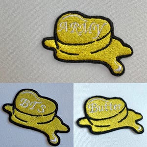 Puede incluir: Tres parches bordados para planchar con forma de barra de mantequilla derritiéndose. Los parches son amarillos con contornos negros. El parche superior dice "ARMY" en letras blancas. El parche del medio dice "BTS" en letras blancas. El parche inferior dice "Butter" en letras blancas.