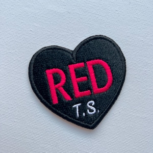 Peut inclure: Patch en forme de cœur noir avec le mot "RED" en lettres roses et les initiales "T.S." en lettres blanches.