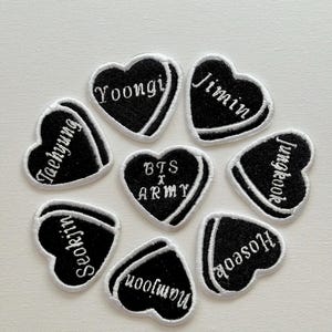 Könnte beinhalten: Acht schwarze herzförmige Patches mit weiß gesticktem Text. Die Patches zeigen die Namen der Mitglieder der südkoreanischen Boyband BTS: Jin, Suga, J-Hope, RM, Jimin, V, Jungkook und den Text "BTS x ARMY".