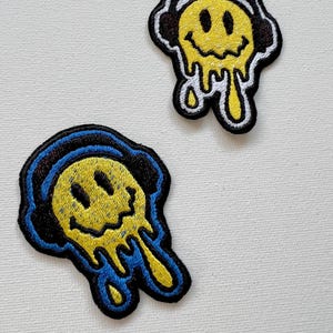 May include: Two embroidered iron-on patches featuring a yellow smiley face with dripping yellow paint and black headphones. One patch has blue trim and the other has white trim.