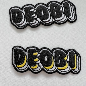May include: Two black embroidered patches with the word "DEOBI" in a stylized, graffiti-like font. The top patch has white outlines, while the bottom patch has yellow outlines.