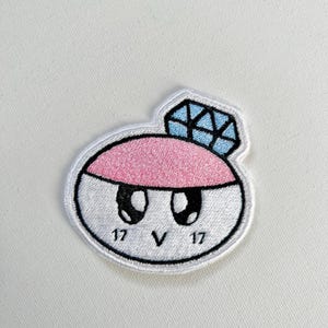 May include: White, pink and blue embroidered patch of a cartoon ring with a diamond on top. The ring has eyes and a mouth, and the text "17 V 17" is written below the ring.