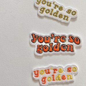 Op de afbeelding: Drie geborduurde patches met de zin "You're so golden" in een retro lettertype. De patches hebben een witte rand en de tekst is goud en oranje. De patches staan op een witte achtergrond.