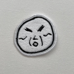 May include: White embroidered patch with a cartoon face expressing frustration. The face has furrowed brows, a mouth open in a gasp, and a single tear coming from the eye.