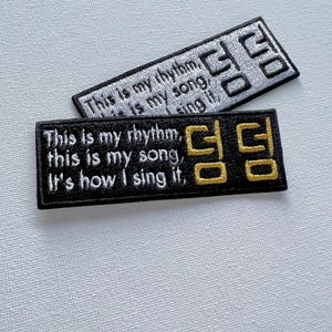 May include: Two black and white embroidered patches with the text "This is my rhythm, this is my song, It's how I sing it." and the Korean word "덤" in gold. The patches are on a white background.