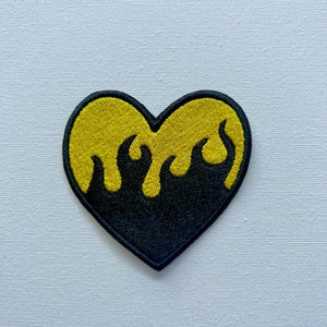 Peut inclure: Un patch brodé en forme de cœur noir et jaune avec un motif de flamme. Le cœur est rempli de jaune et les flammes sont noires.