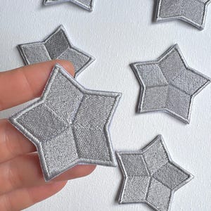 Könnte beinhalten: Sechs silberne, sternförmige Bügelbilder mit einem karierten Muster. Die Patches befinden sich auf einem weißen Hintergrund.