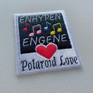 Peut inclure: Un patch brodé blanc avec un design de cadre photo noir et blanc. Le cadre contient le texte "ENHYPEN ENGENE" et des notes de musique en rose, bleu, jaune et vert. Un cœur rouge se trouve dans le coin inférieur gauche du cadre. Le texte "Polaroid Love" est brodé sous le cadre.