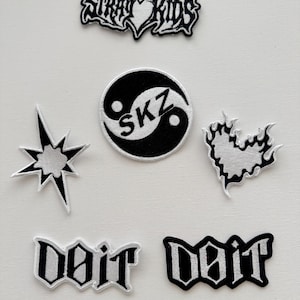 以下が含まれることがあります： 白黒の刺繍パッチのコレクション。デザインには、「Stray Kids」の文字、SKZの陰陽シンボル、スターバースト、ハート型の炎、ゴシックフォントの「Doit」という単語が含まれています。