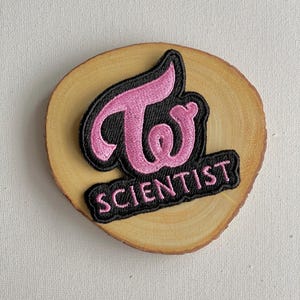 May include: A pink embroidered patch with the word "Twice" in a stylized font, above the word "Scientist". The patch has a black border and is set on a round, light brown wooden slice.