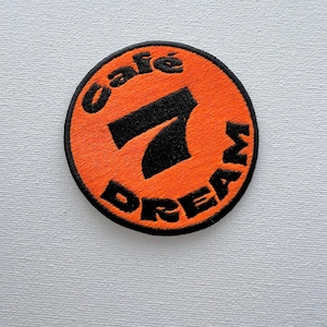 Könnte beinhalten: Orangefarbener und schwarzer gestickter Aufnäher mit dem Text "Cafe 7 Dream" in einem Kreis.