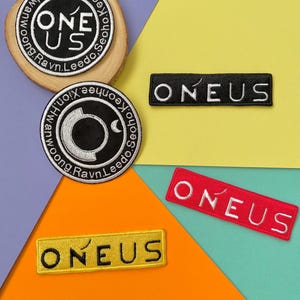 May include: Four embroidered patches featuring the band name "ONEUS" in different colors and styles. The patches are on a colorful background.