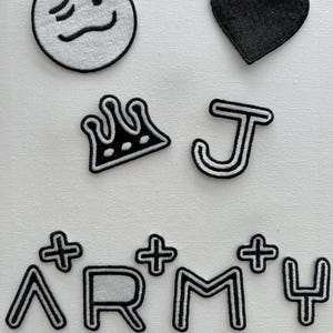 May include: A set of iron-on patches featuring a black heart, a crown, a letter J, a white smiley face, and the word "ARMY" with plus signs on either side of the letters.