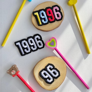 Könnte beinhalten: Vier gestickte Patches mit den Zahlen 1996 und 96 auf weißem Hintergrund. Die Patches werden auf Holzrundlingen mit bunten Bleistiften und Radiergummis präsentiert.