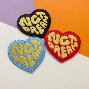 Peut inclure: Trois patchs en forme de cœur avec le texte "NOT DREAMS" en lettres jaunes et cursive. Les patchs sont sur un fond violet, blanc et orange. Les patchs sont noirs, rouges et bleu clair.