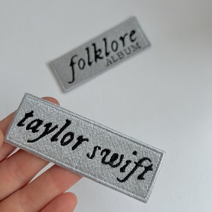 Op de afbeelding: Twee grijze stoffen patches met zwarte geborduurde tekst. Eén patch zegt "folklore ALBUM" en de andere zegt "taylor swift".