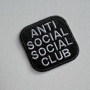 Puede incluir: Parche cuadrado negro con texto bordado en blanco que dice "ANTI SOCIAL SOCIAL CLUB".