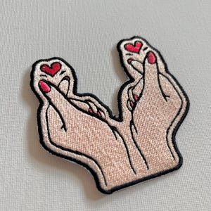 Peut inclure: Écusson brodé représentant deux mains formant un cœur. Les mains sont de couleur claire avec du vernis à ongles rouge et de petits cœurs rouges. L'écusson a une bordure noire, idéal pour les vêtements ou les accessoires.