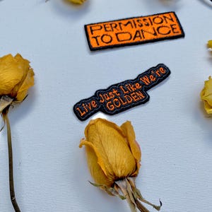 May include: Two iron-on patches with orange text on a black background. The top patch says "Permission to Dance" and the bottom patch says "Live Just Like We're Golden". The patches are on a white surface with dried yellow roses.