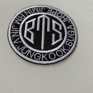 Pode incluir: Um remendo bordado em preto e branco com as letras "BTS" em um círculo. O círculo é cercado pelos nomes dos membros da banda: "JUNGKOOK, SUGA, J-HOPE, JIMIN, V, JIN, RM".